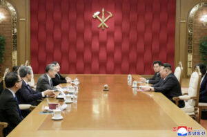 diplomatic meeting in a wood floored conference room with officials seated across a long table a red padded wall behind them and a gold emblem above